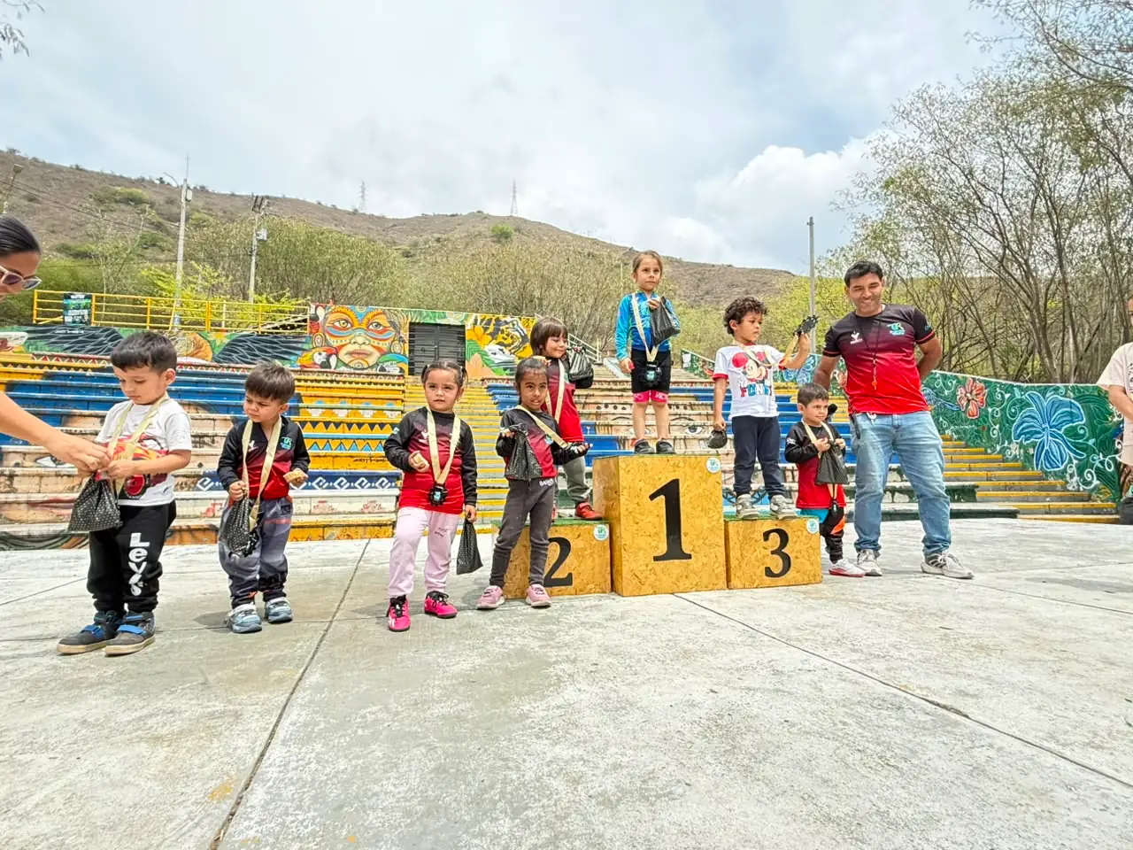 Una decena de niños de las categorías más pequeñas posan con sus medallas y sus bolsas de premio junto al podio, acompañados por dos adultos del Club Trocha y Ruta