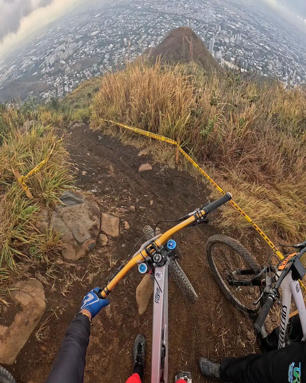 Vista desde el pecho de un corredor, sobre el manubrio: el sendero marcado con cinta amarilla en lo alto del cerro, con la ciudad de Cali abajo, Cali Bike Park, 2026