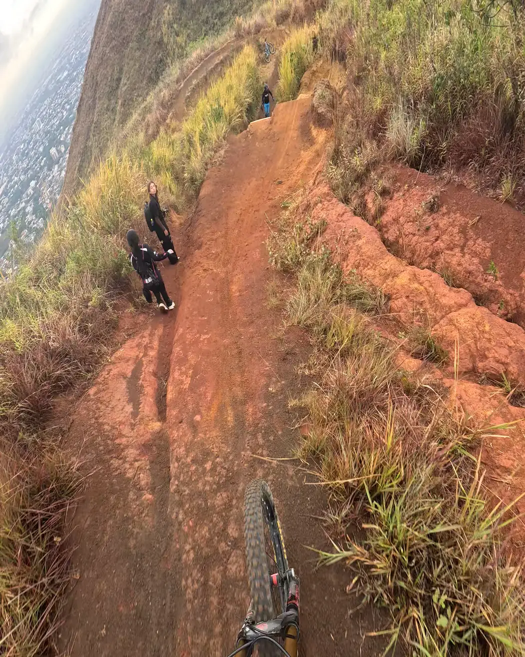Vista desde el manubrio: un tramo de tierra roja baja por la ladera, con otras personas más adelante, Cali Bike Park, 2026